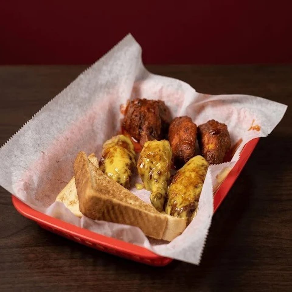 Red basket lined with paper holds grilled kebabs, fried meat, toasted bread, and roasted vegetables on a wooden table.
