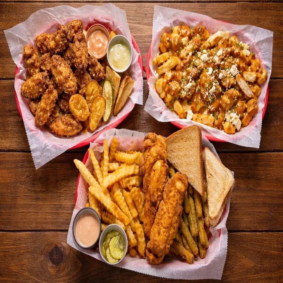 Three baskets of fried chicken, loaded fries, crinkle-cut fries, pickles, toast, and dipping sauces arranged on a wooden table.